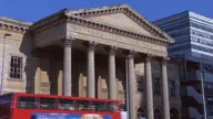 A neoclassical building with tall columns and a triangular pediment stands beside a modern office block. A red double-decker bus passes by in the foreground under a clear blue sky.