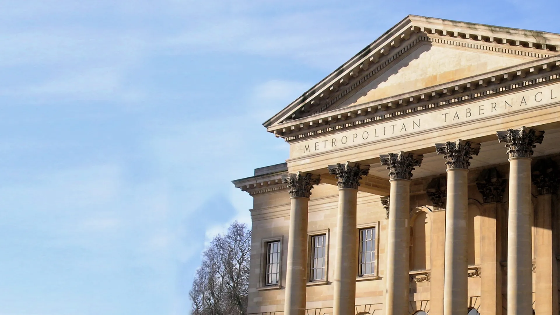 A neoclassical building with tall columns and a triangular pediment inscribed “Metropolitan Tabernacle” stands under a clear blue sky. Trees are visible in the background.