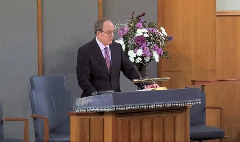 Man in suit speaking at a podium with floral arrangement in a formal setting.