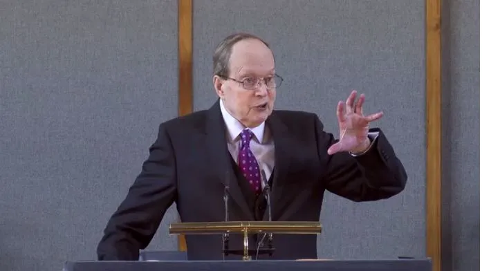 Elderly man in a suit giving a speech at a podium with a microphone.