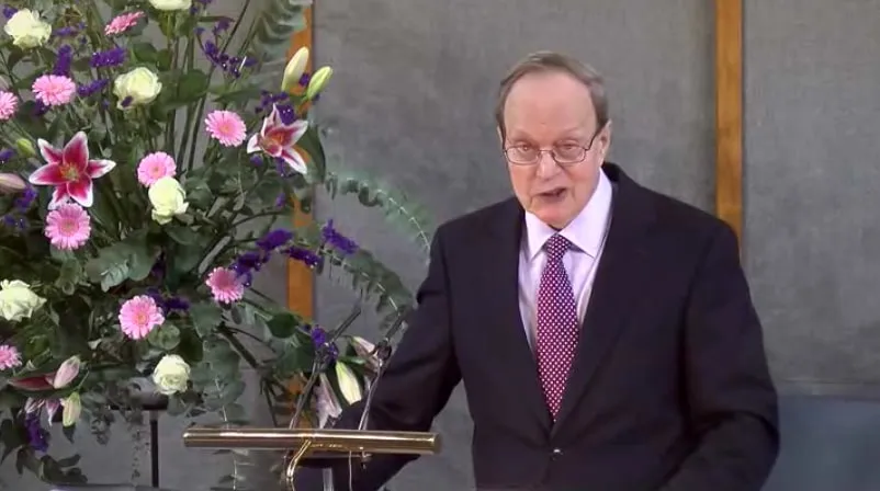 Man speaking at a podium with a large bouquet of pink and white flowers on the left.
